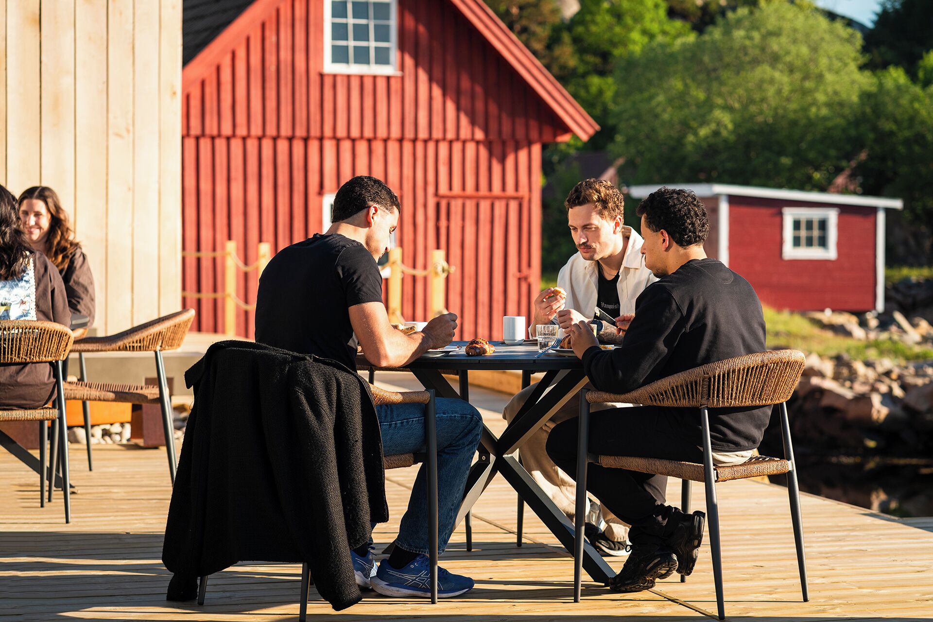 Un groupe de personnes est assis à une table en bois, à l’extérieur, et prend son repas. Des chaises sont disposées autour de la table. On aperçoit, en arrière-plan, un bâtiment rouge et une terrasse en bois.