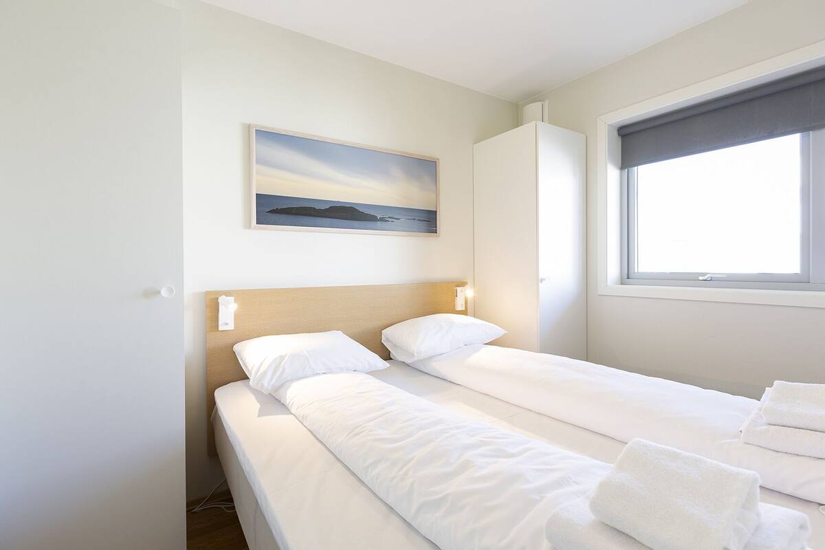 A single bed with white linens, a nightstand, a wall-mounted light, a painting, and a window with a view.