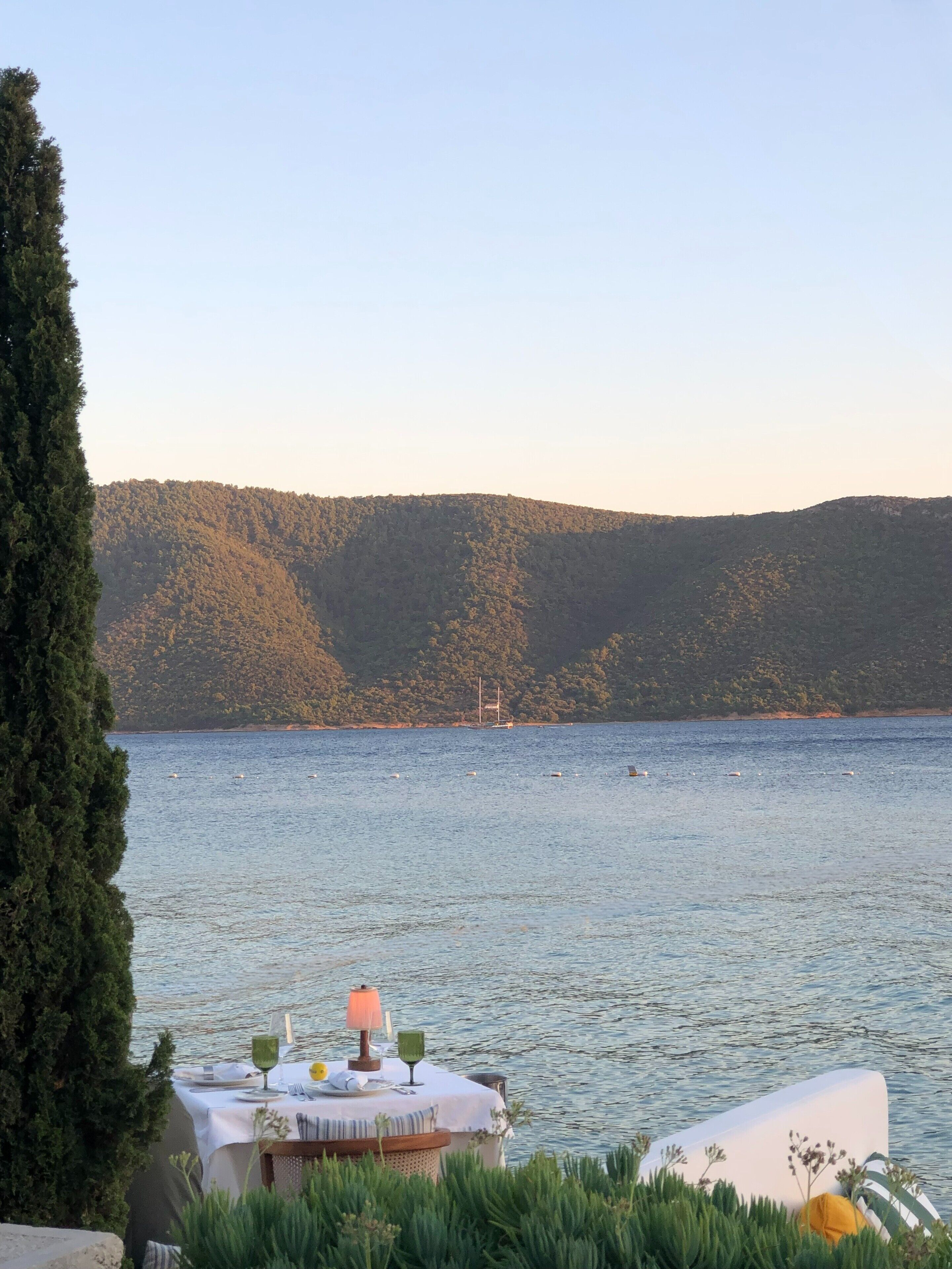 A lakeside table set with a tablecloth, a lamp, and glassware, overlooking a calm lake with a sailboat in the distance and a forested hill in the background.