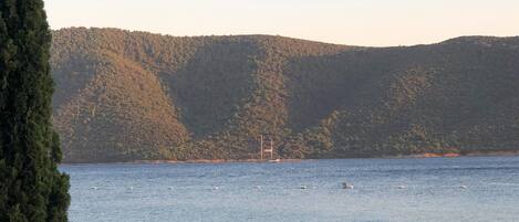 A lakeside table set with a tablecloth, a lamp, and glassware, overlooking a calm lake with a sailboat in the distance and a forested hill in the background.