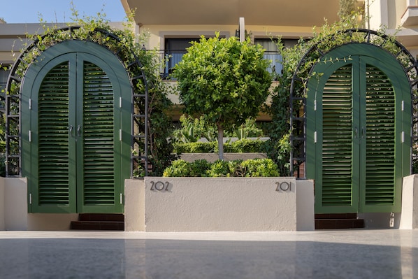 Entrada de edificio con dos puertas arqueadas verdes, un árbol y un pequeño jardín.