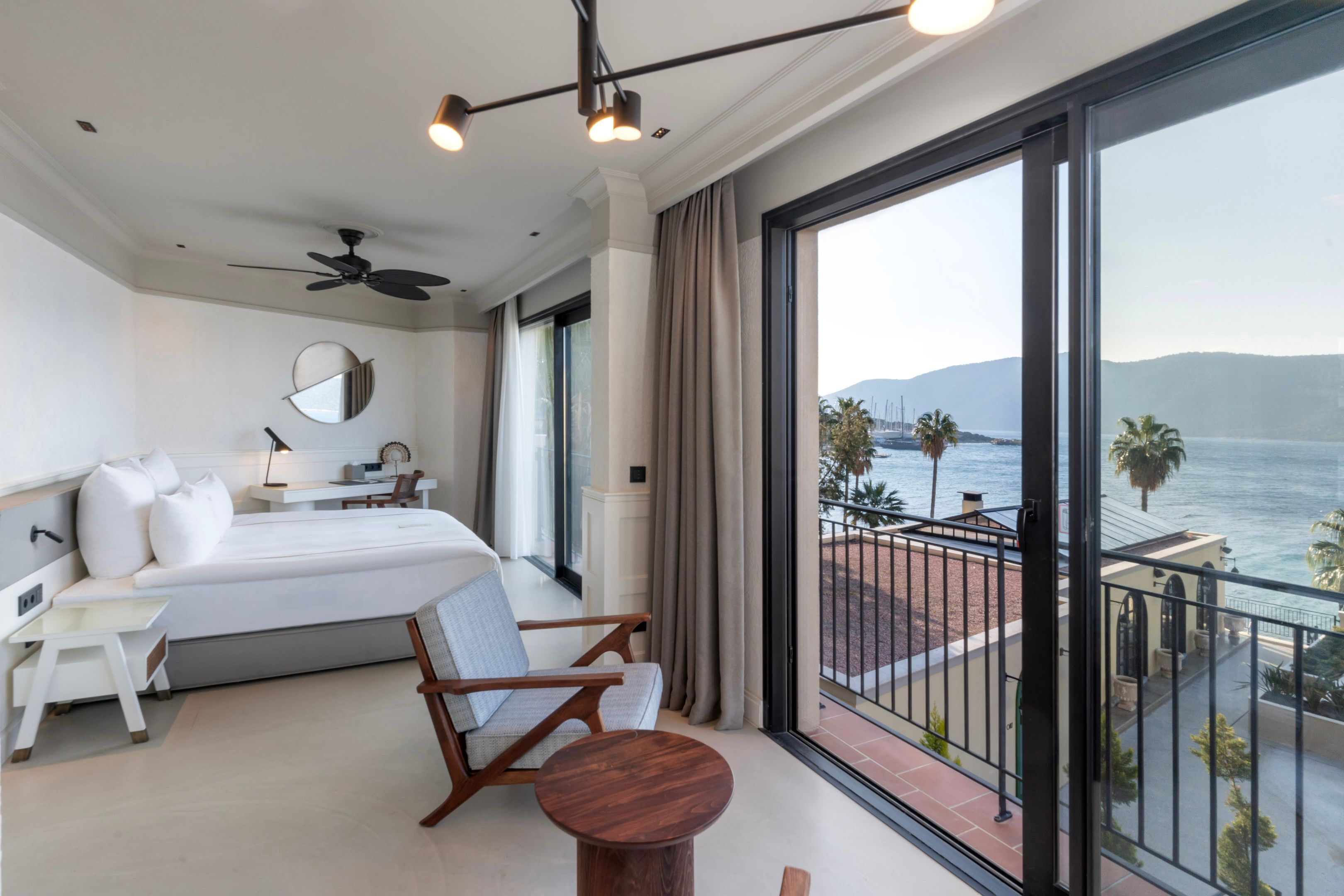 A modern hotel room with a large bed, a desk, a chair, and a balcony with a view of the sea.