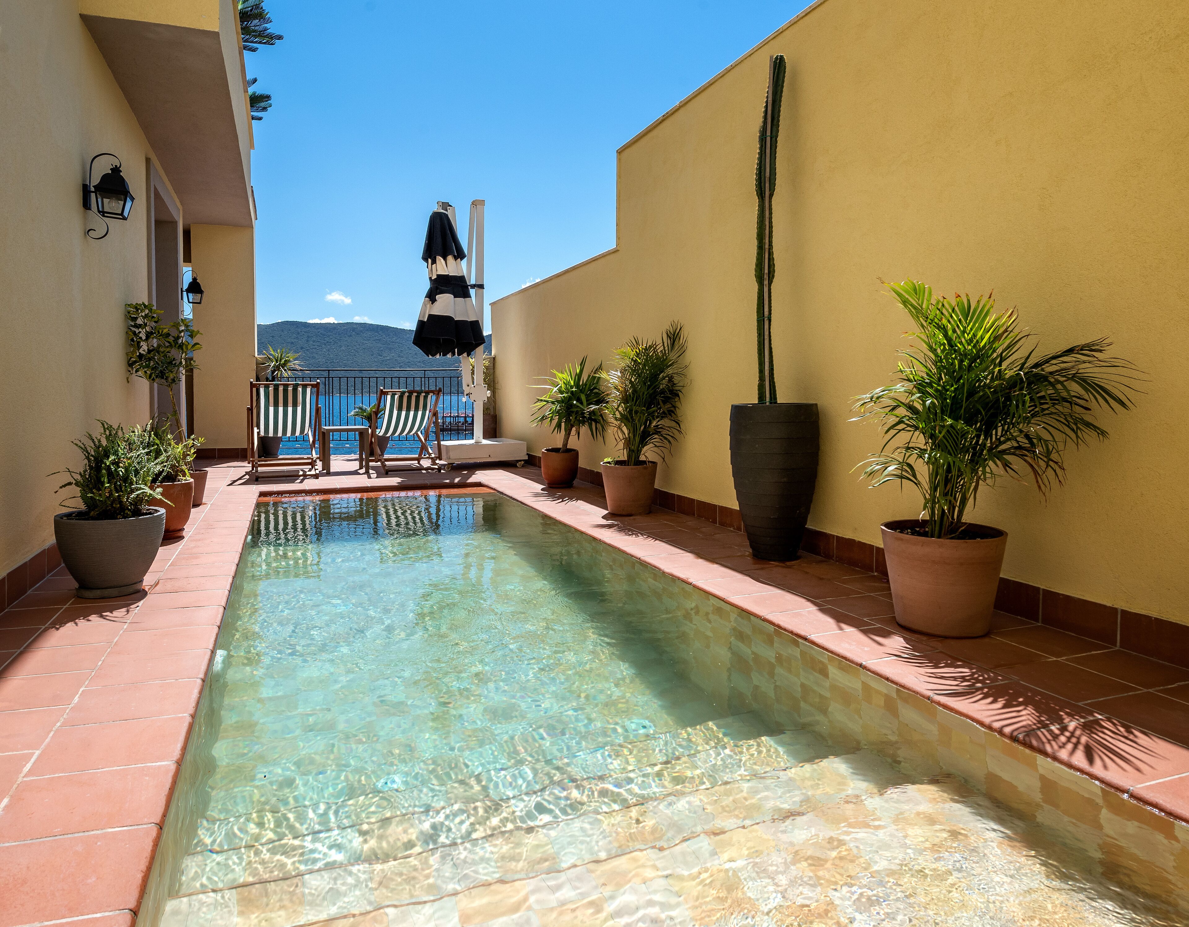 A clear pool with tiled edges, surrounded by potted plants and a chair with a striped cushion.