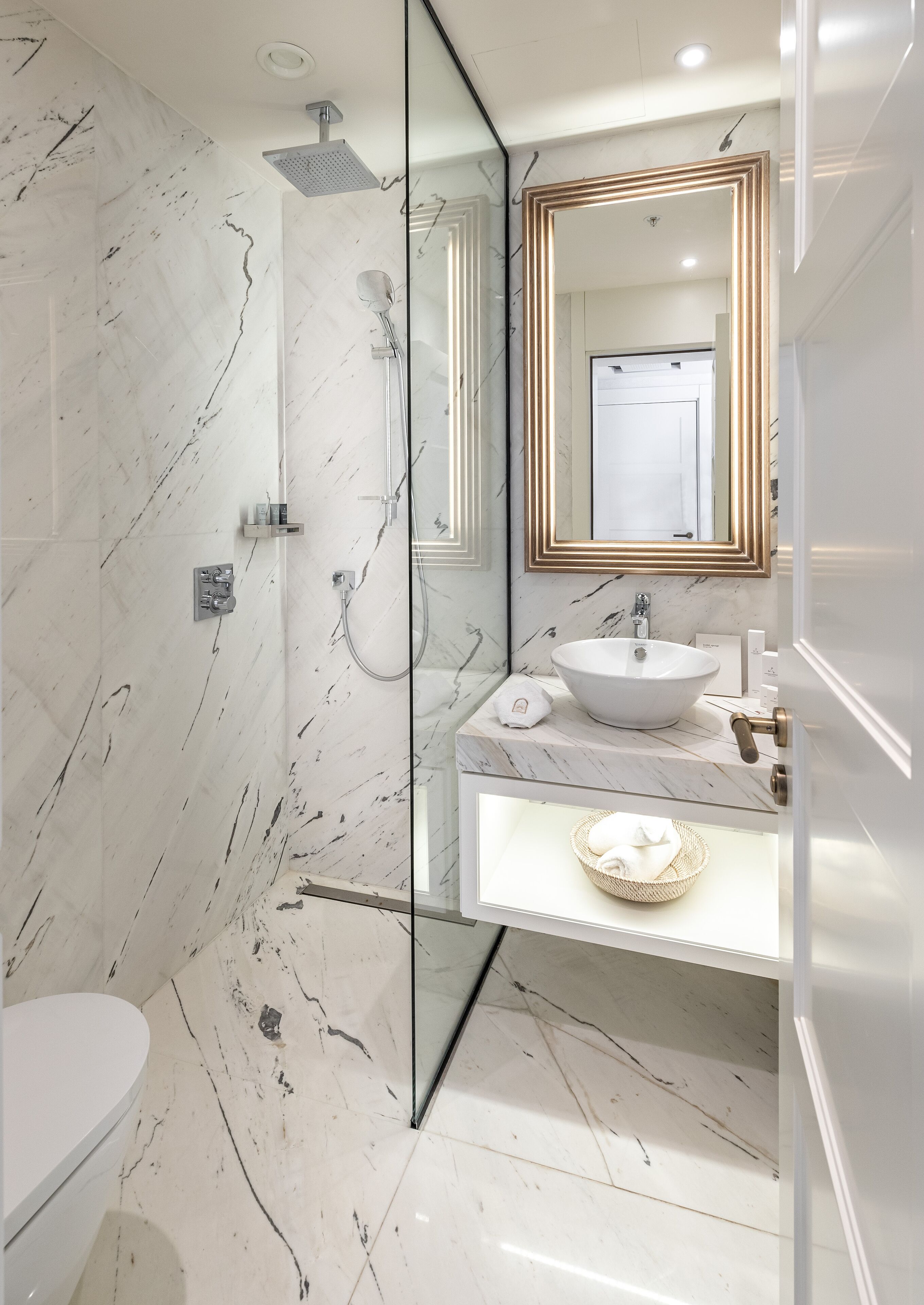 Een moderne badkamer met een douchecabine van glas, een wit marmeren wastafel en een grote spiegel met gouden lijst.