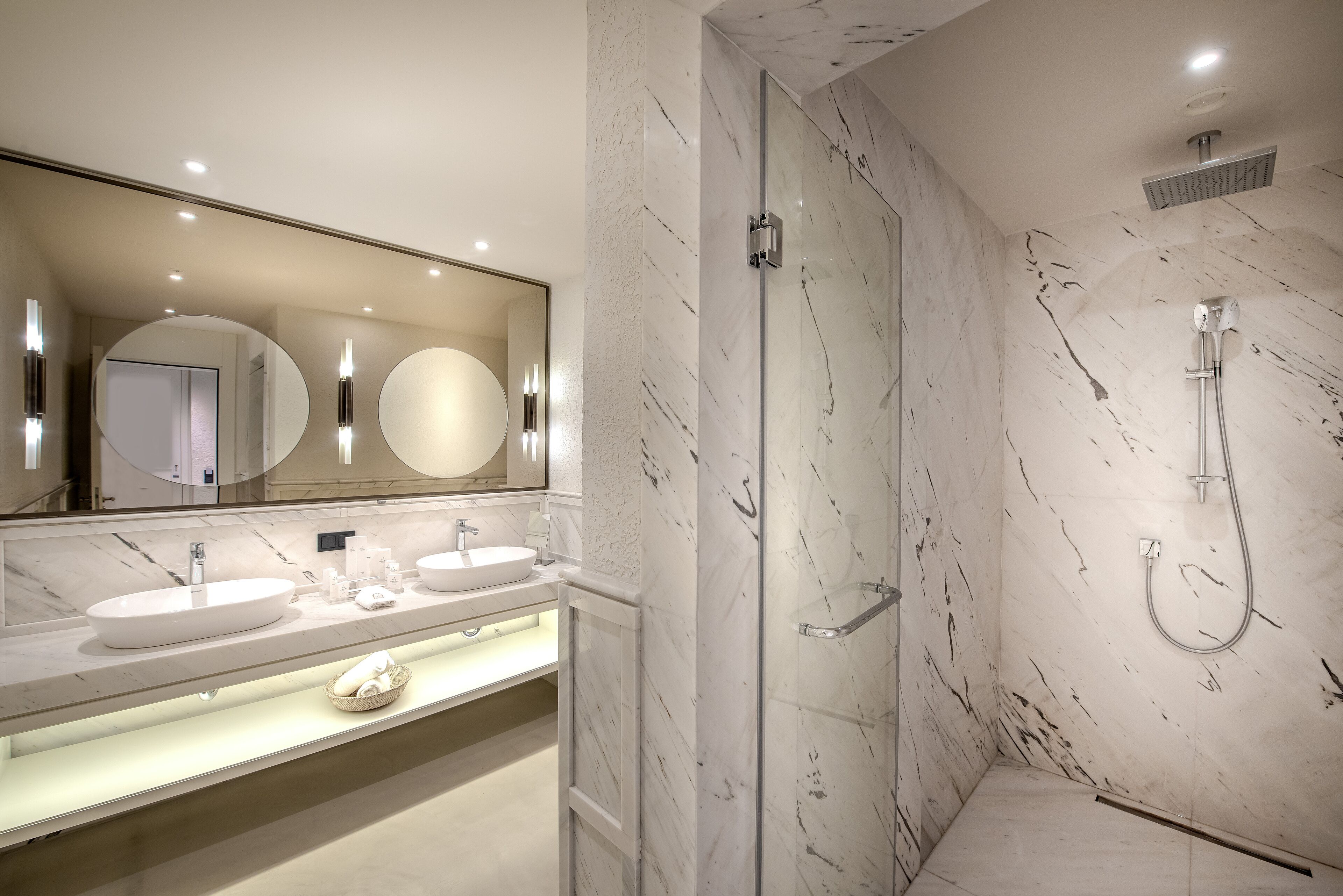 Un baño moderno con un espejo grande, dos lavamanos, una ducha con cerramiento de vidrio y paredes de mármol.