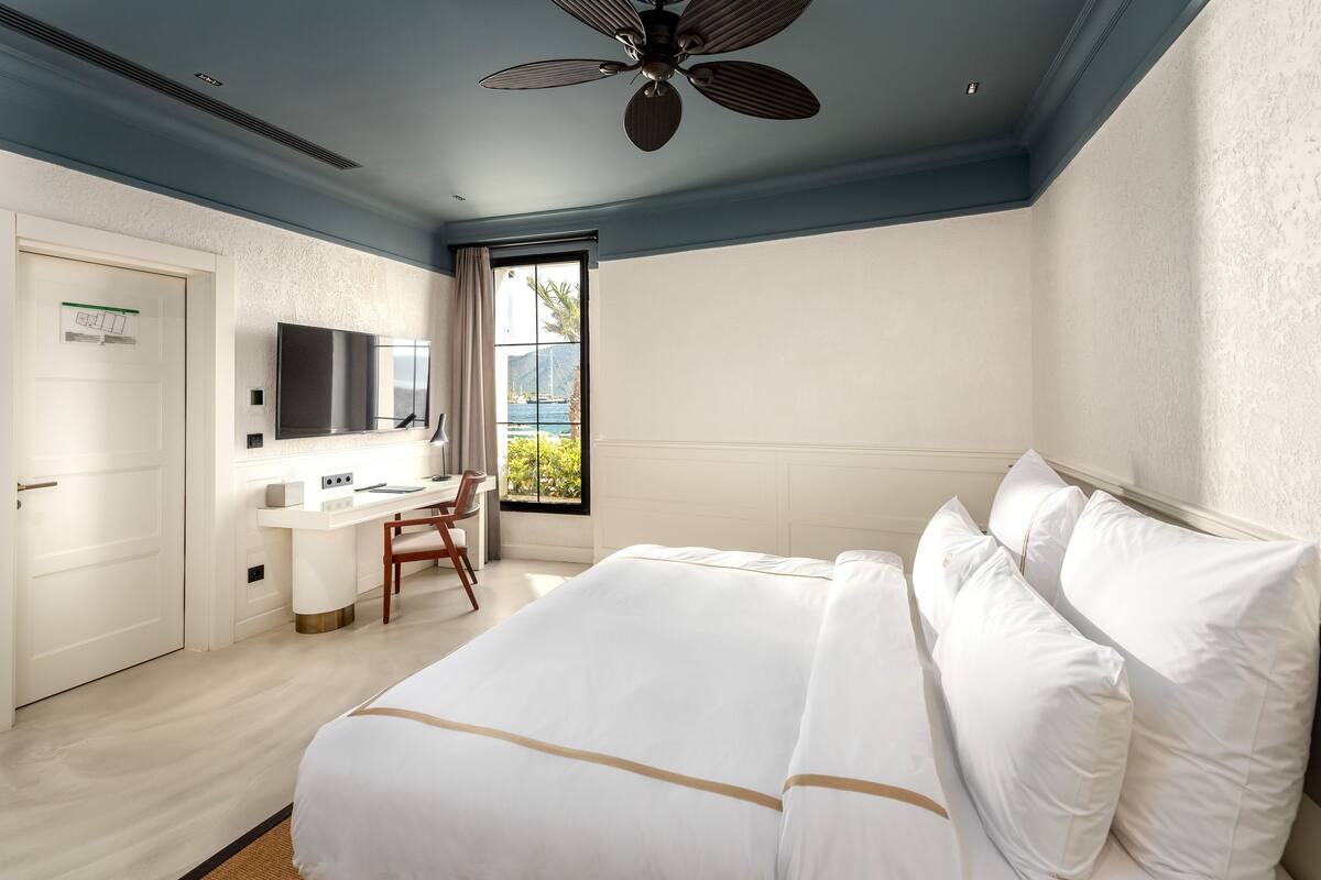 A hotel room with a bed, a desk, a chair, a TV, and a window with a view of greenery.