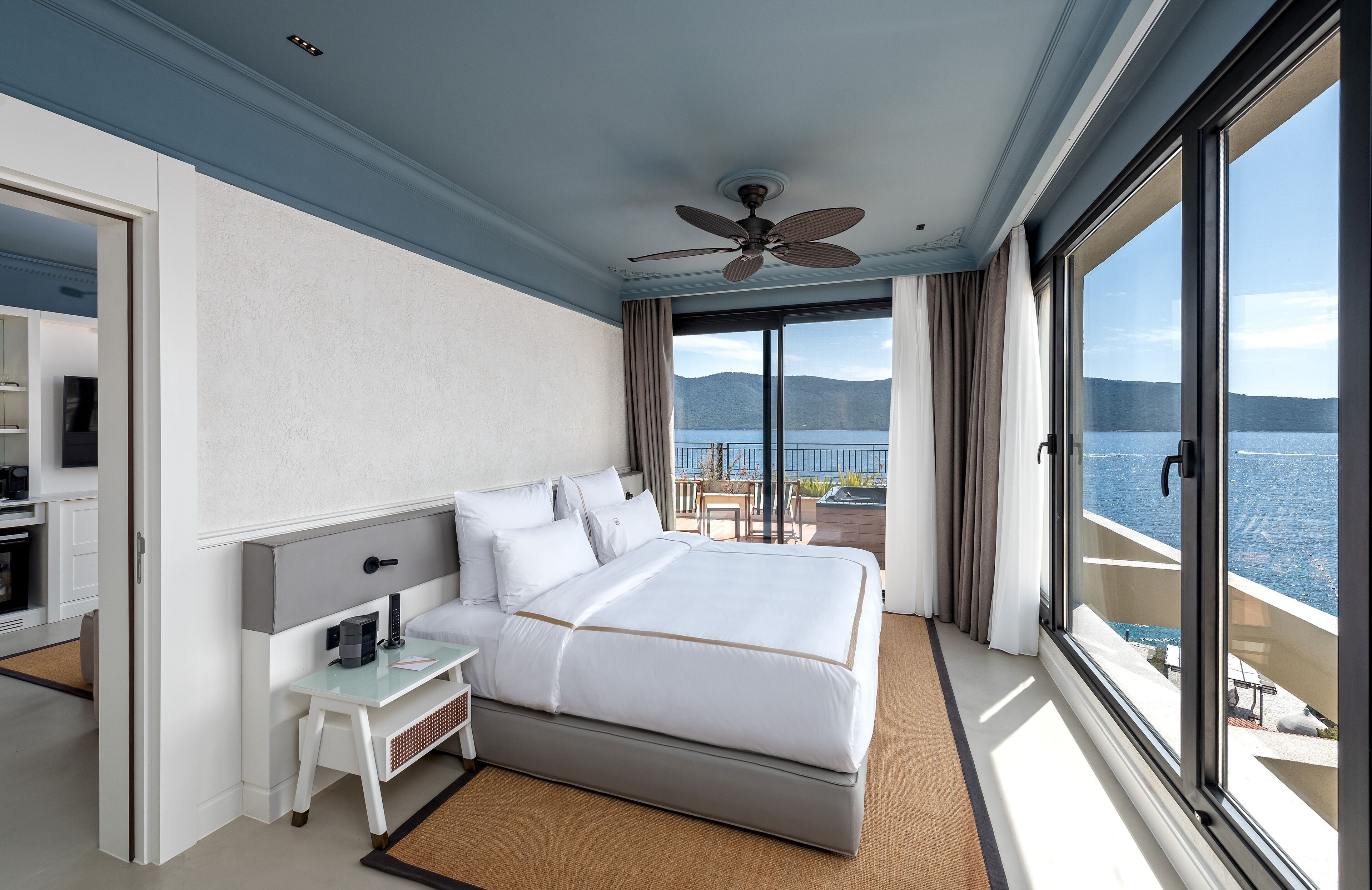 A modern hotel room with a large bed, a desk, and a balcony with a view of the sea.