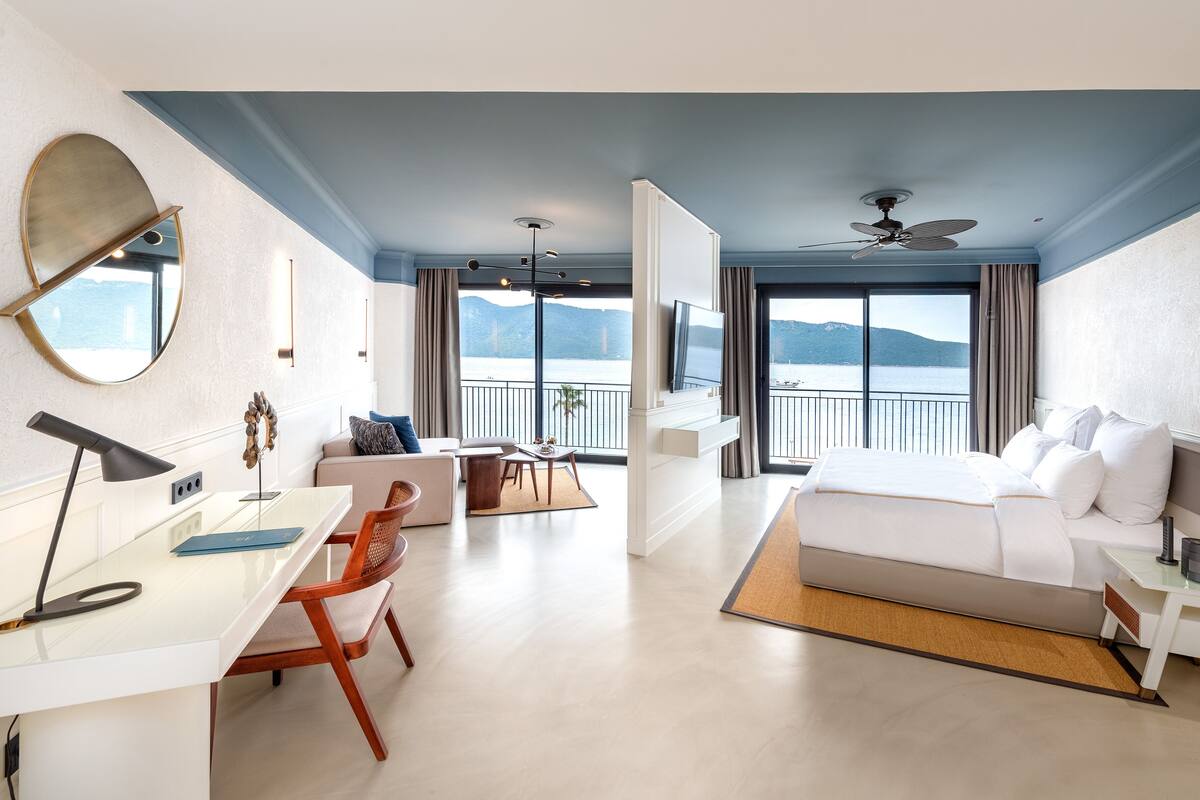 A modern hotel room with a balcony, a bed, a desk, and a seating area.