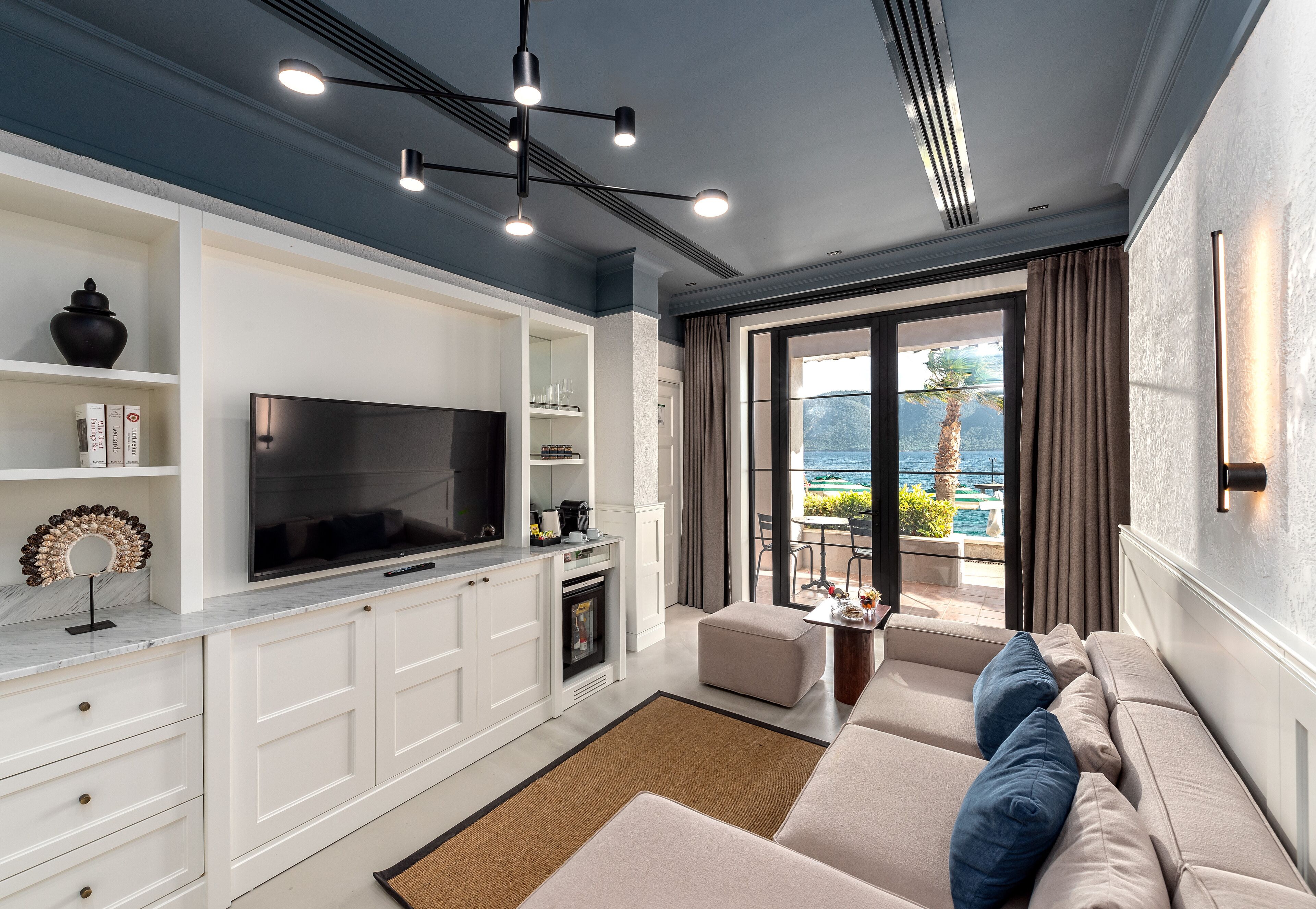 A modern living room with a flat-screen TV, white cabinetry, a sofa with blue cushions, and a view of the ocean through large windows.