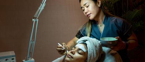 A person receiving a facial treatment under a lamp in a room with a plant and a medical device on a counter.