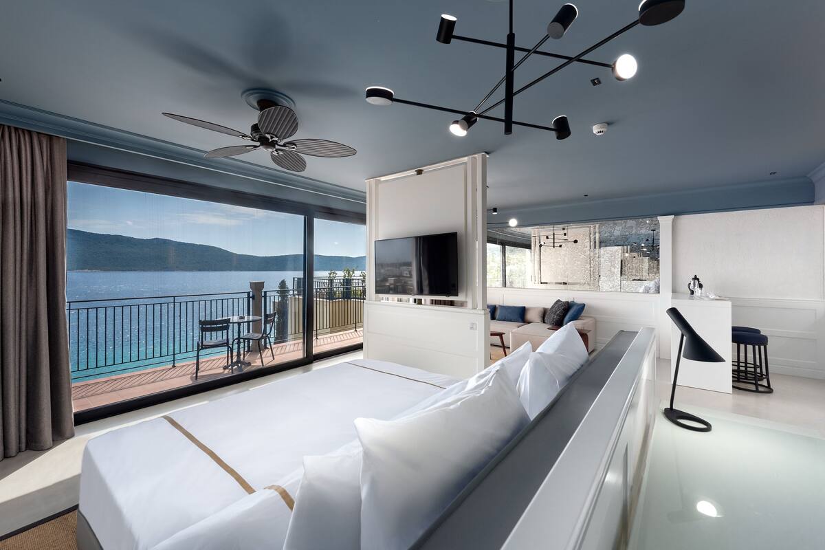 A modern hotel room with a large bed, a TV, a balcony with a view of the sea, and a dining area.