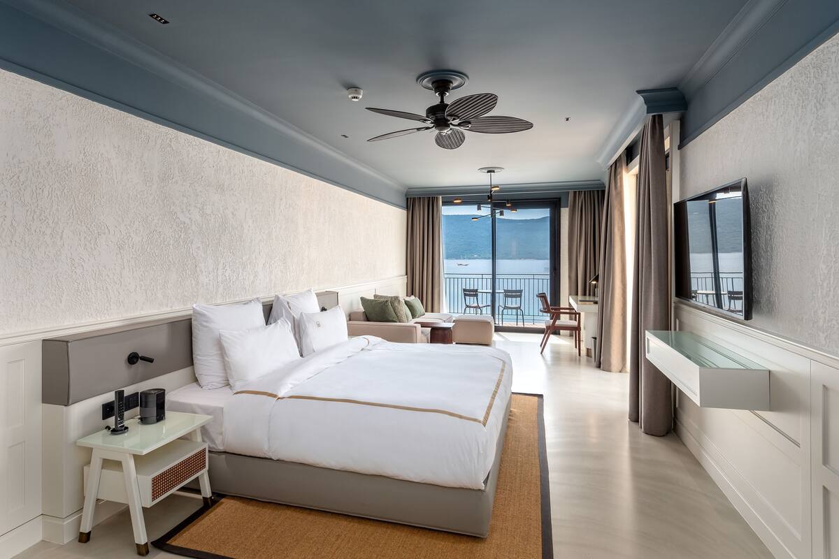 A modern hotel room with a large bed, a desk, a chair, and a balcony with a view.