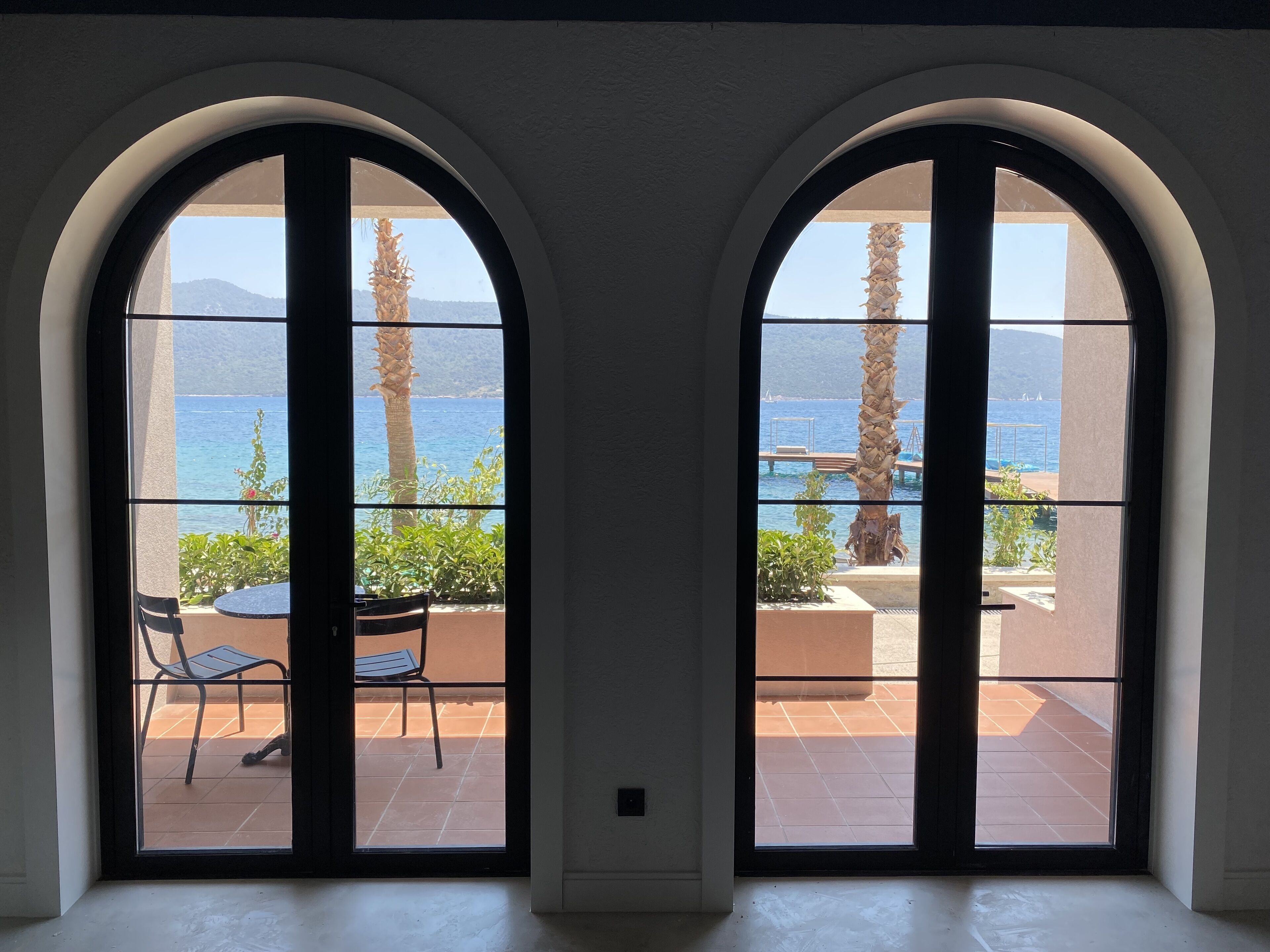 A room with two large arched windows overlooking a terrace with a table, chairs, and a palm tree.