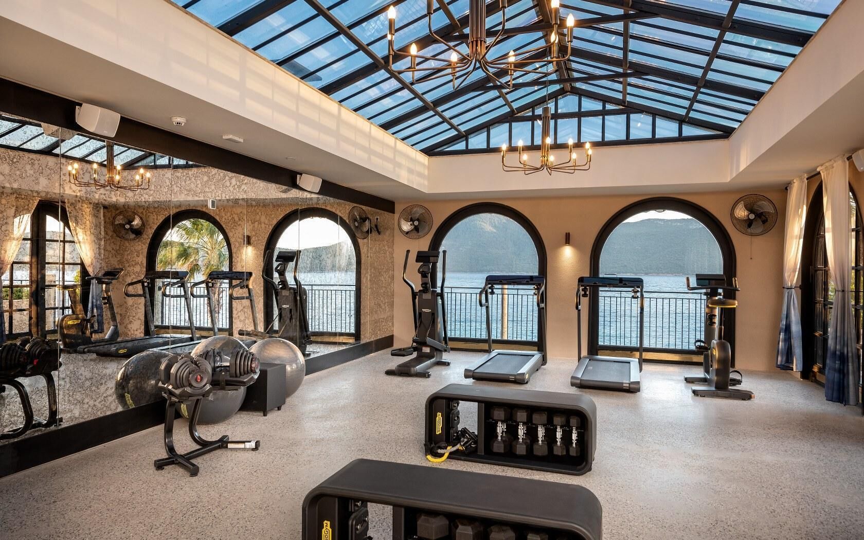 A gym with exercise equipment, large windows, and a glass ceiling overlooking a body of water.