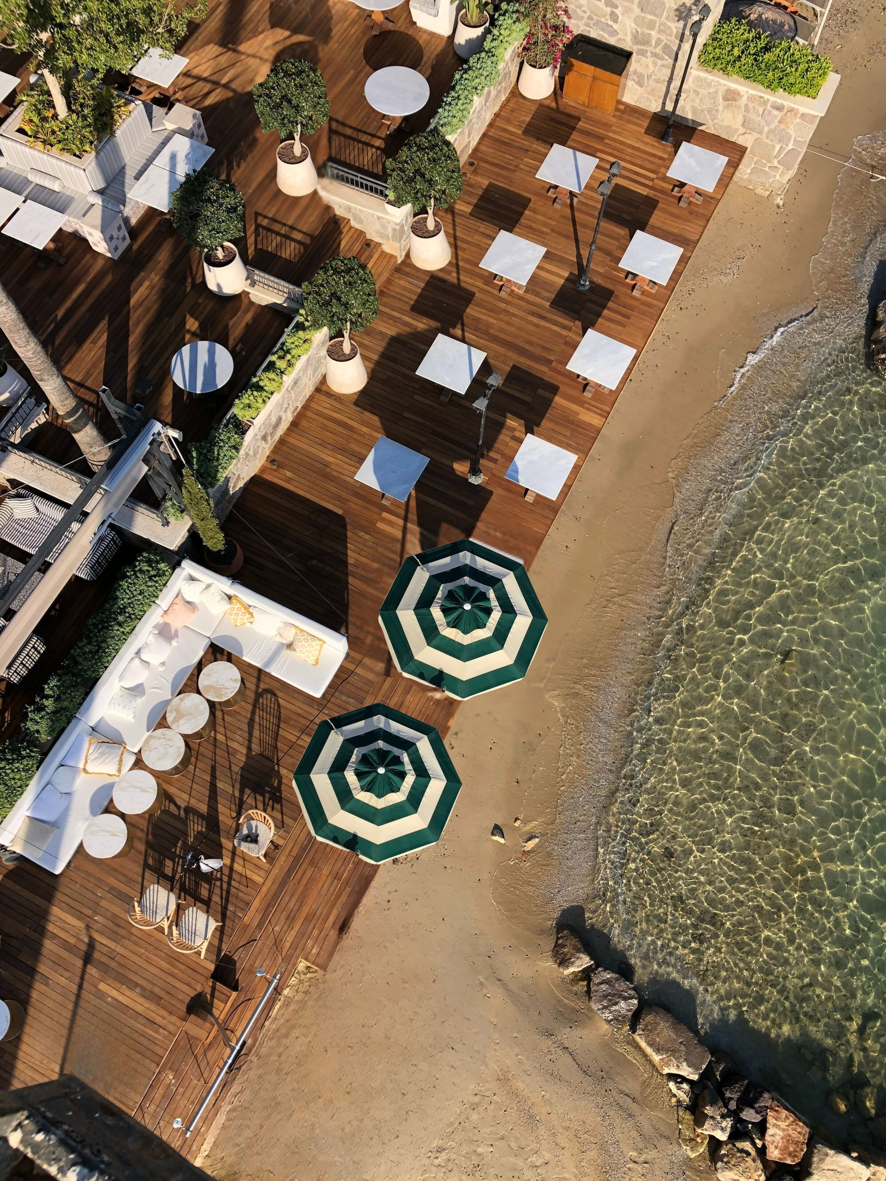 An aerial view of a beachside patio with wooden flooring, white round tables, and green umbrellas.