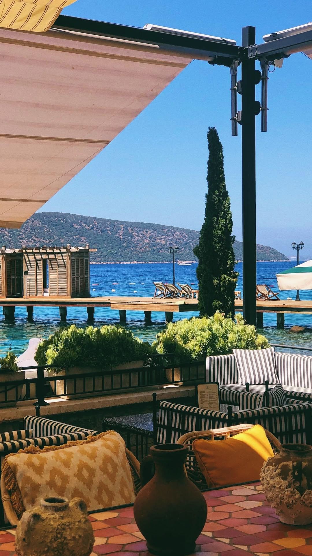 Un espacio de estar al aire libre con vista al mar, muebles de madera y una pérgola.