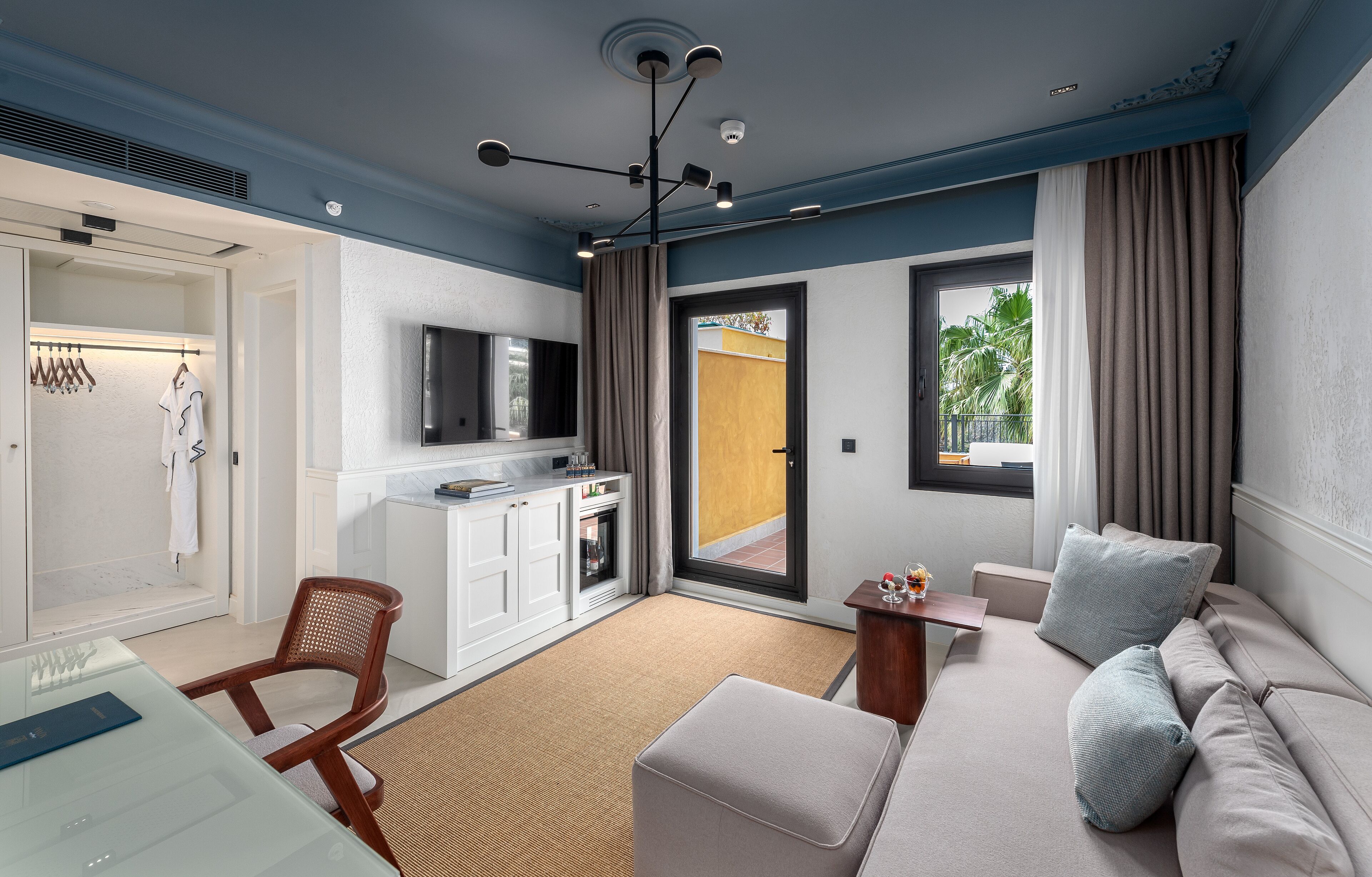 A modern hotel room with a sofa, a desk, a television, and a sliding door to the balcony.