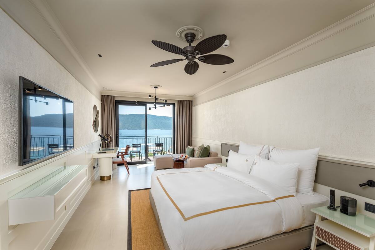 A hotel room with a large bed, a ceiling fan, a TV, a balcony with a view of the sea, and a desk with a chair.