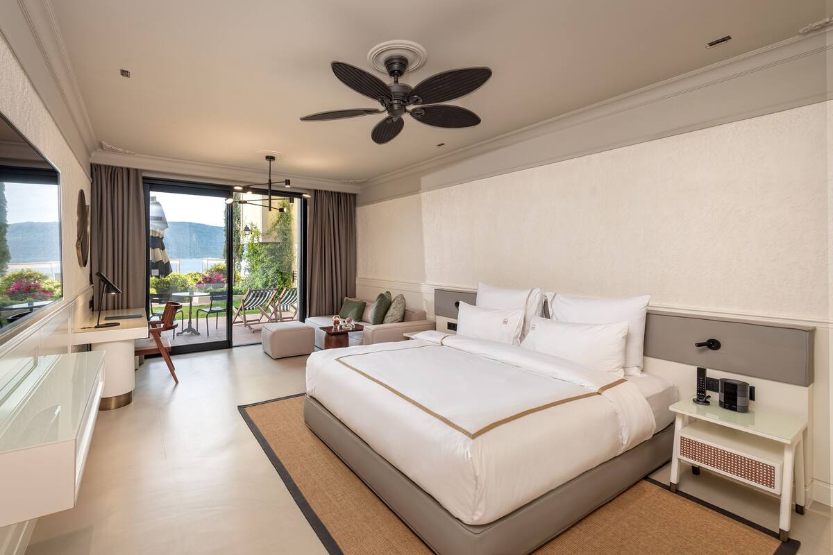 A modern hotel room with a large bed, a desk, a chair, a ceiling fan, and a view of the outdoors.