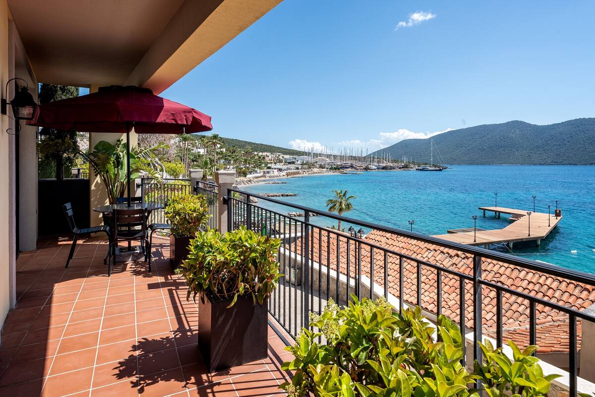 A balcony with a red umbrella, potted plants, and a view of the sea and marina.