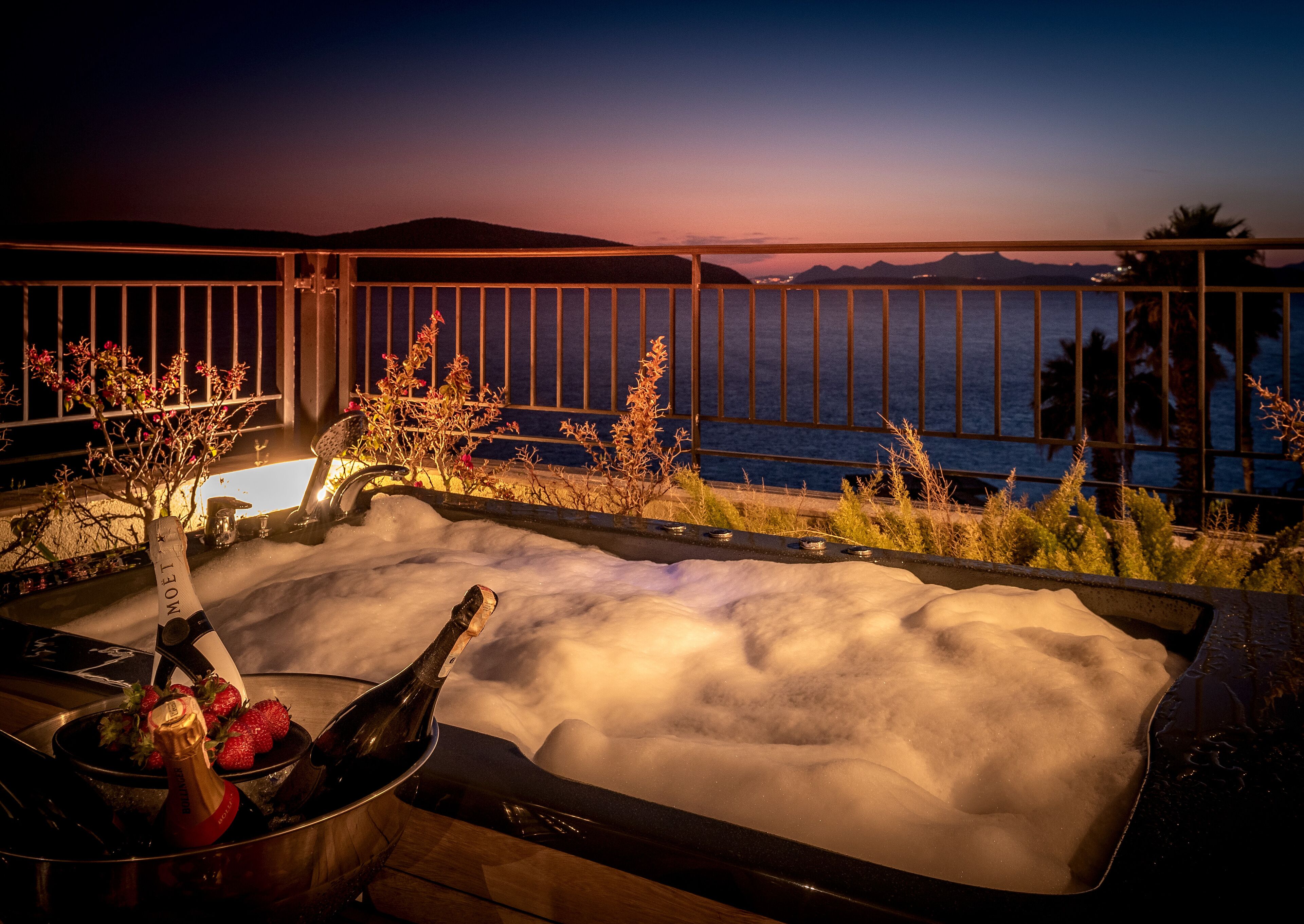 A hot tub with champagne and strawberries, overlooking a body of water and mountains at sunset.