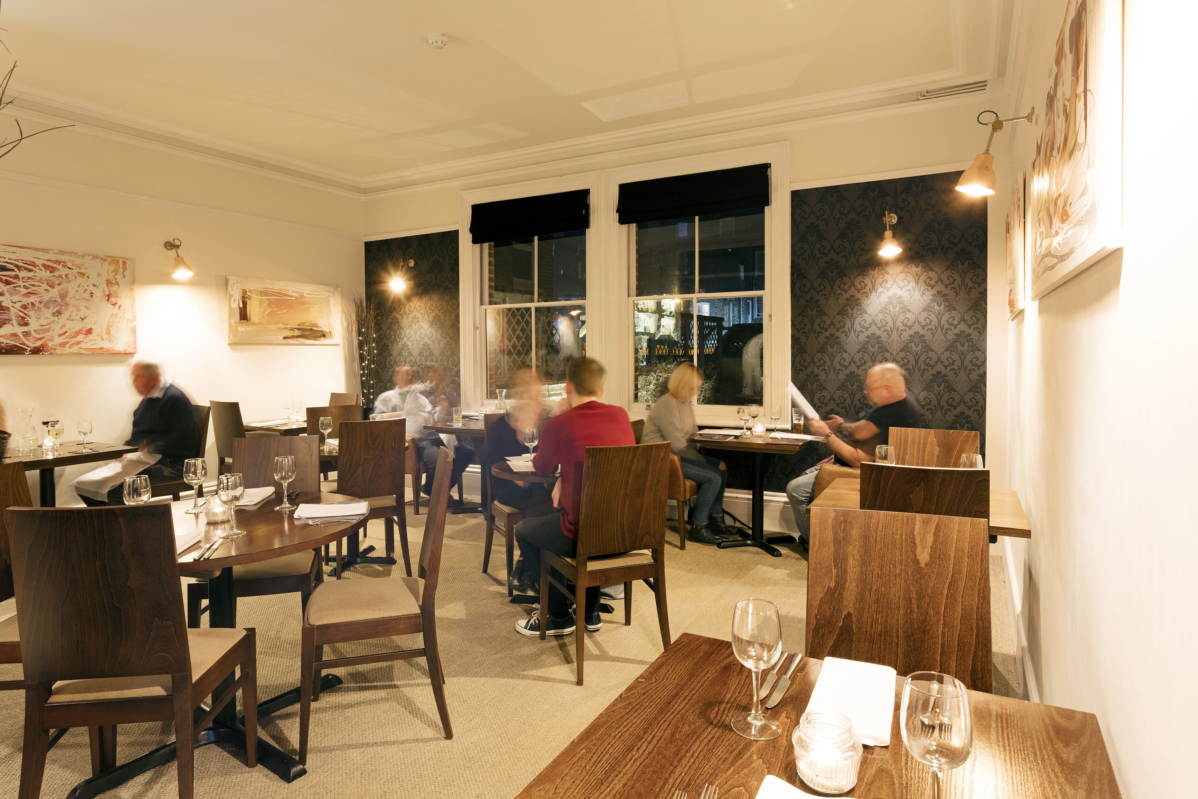 Un restaurant amb taules i caires de fusta, finestres grans amb cortines negres i obra d'art a les parets.