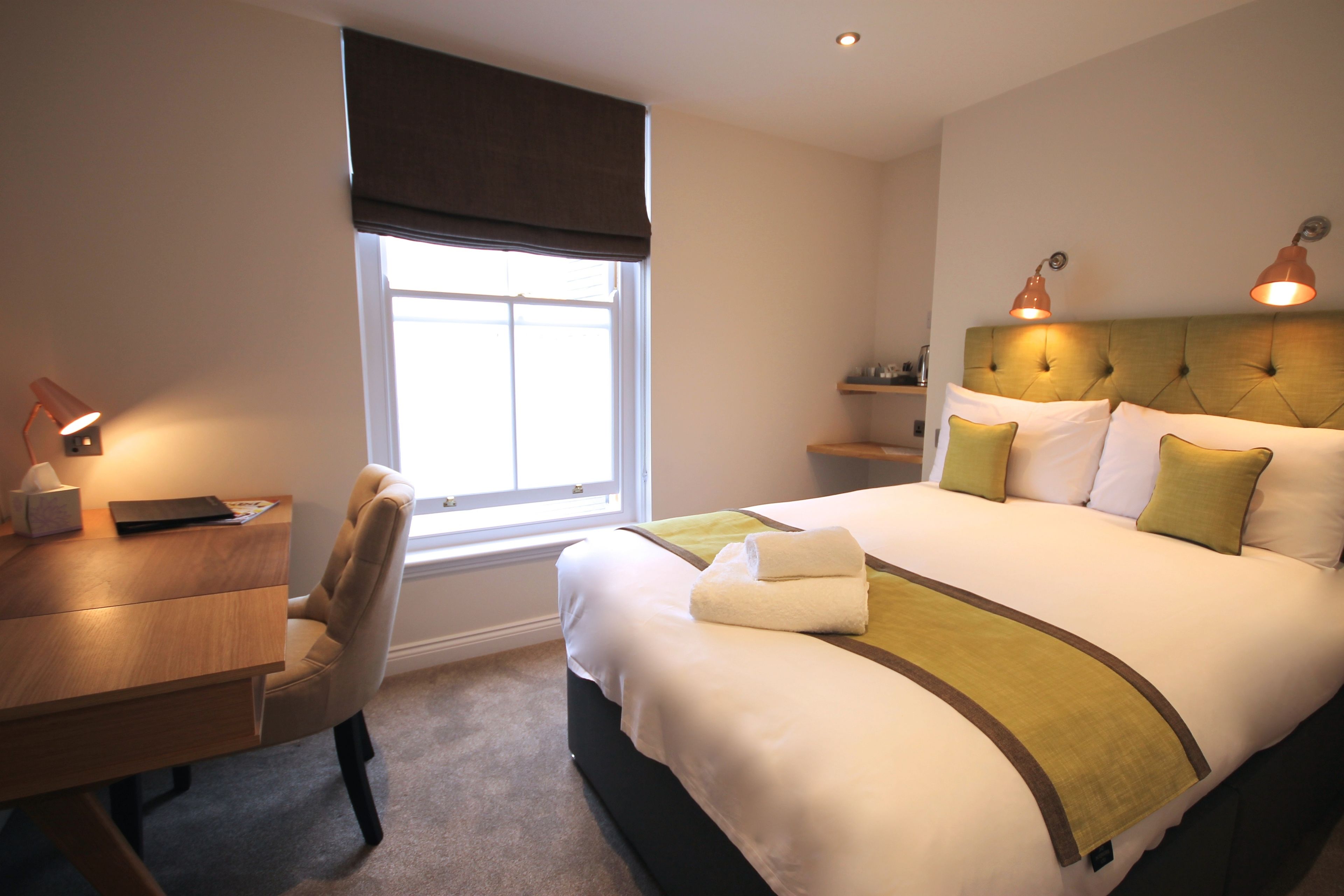 A hotel room with a bed, a desk, a chair, a window with a view, and a nightstand with a lamp.