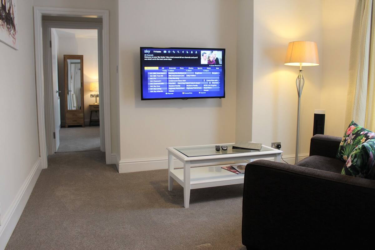 Een woonkamer met een flatscreen televisie aan de muur, een salontafel met glazen blad, een bank en een vloerlamp.