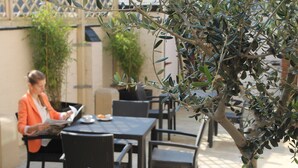 Una persona leyendo una revista en una mesa al aire libre con sillas y una planta en maceta cerca.
