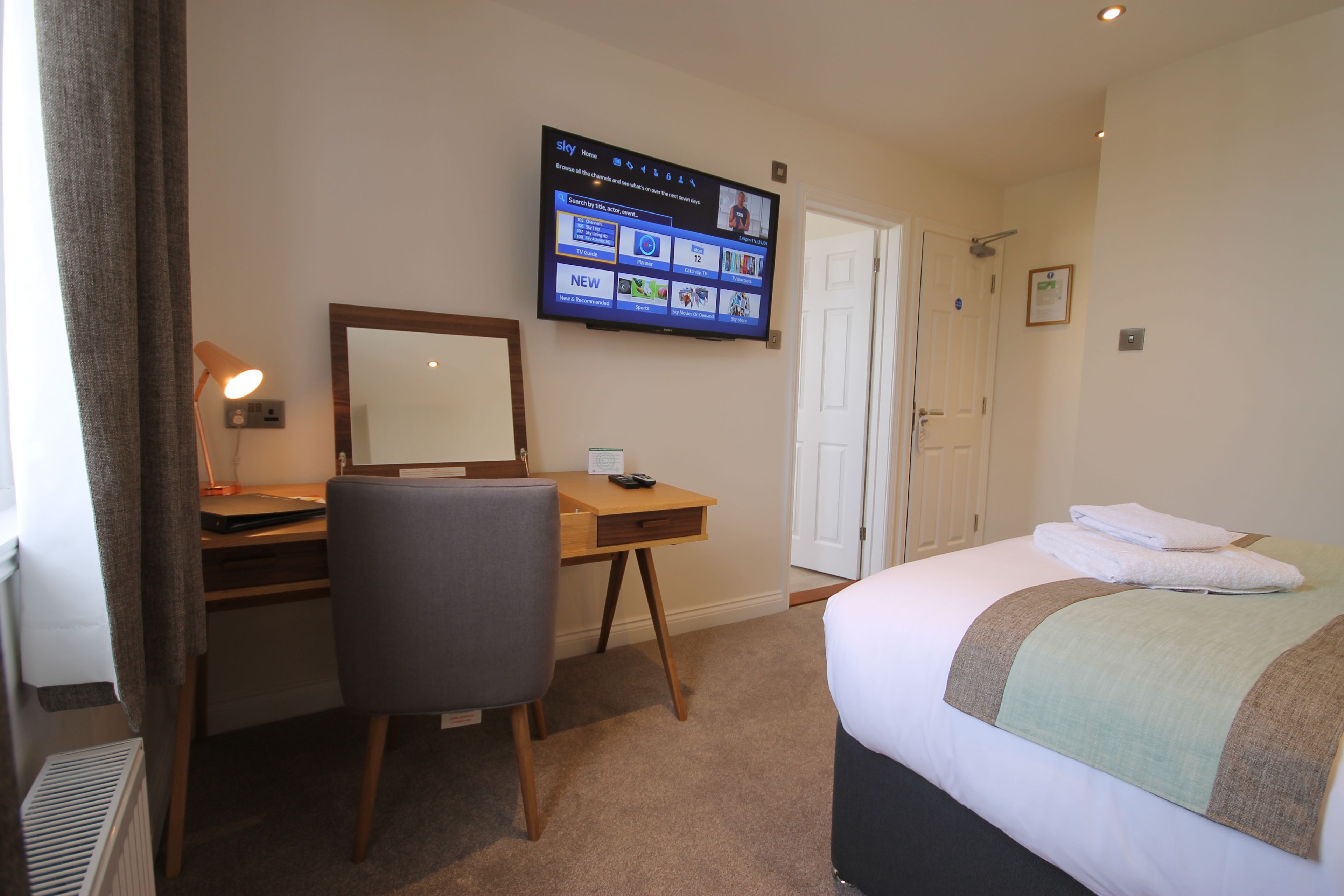 A hotel room with a bed, a desk, a chair, a TV mounted on the wall, and a door.