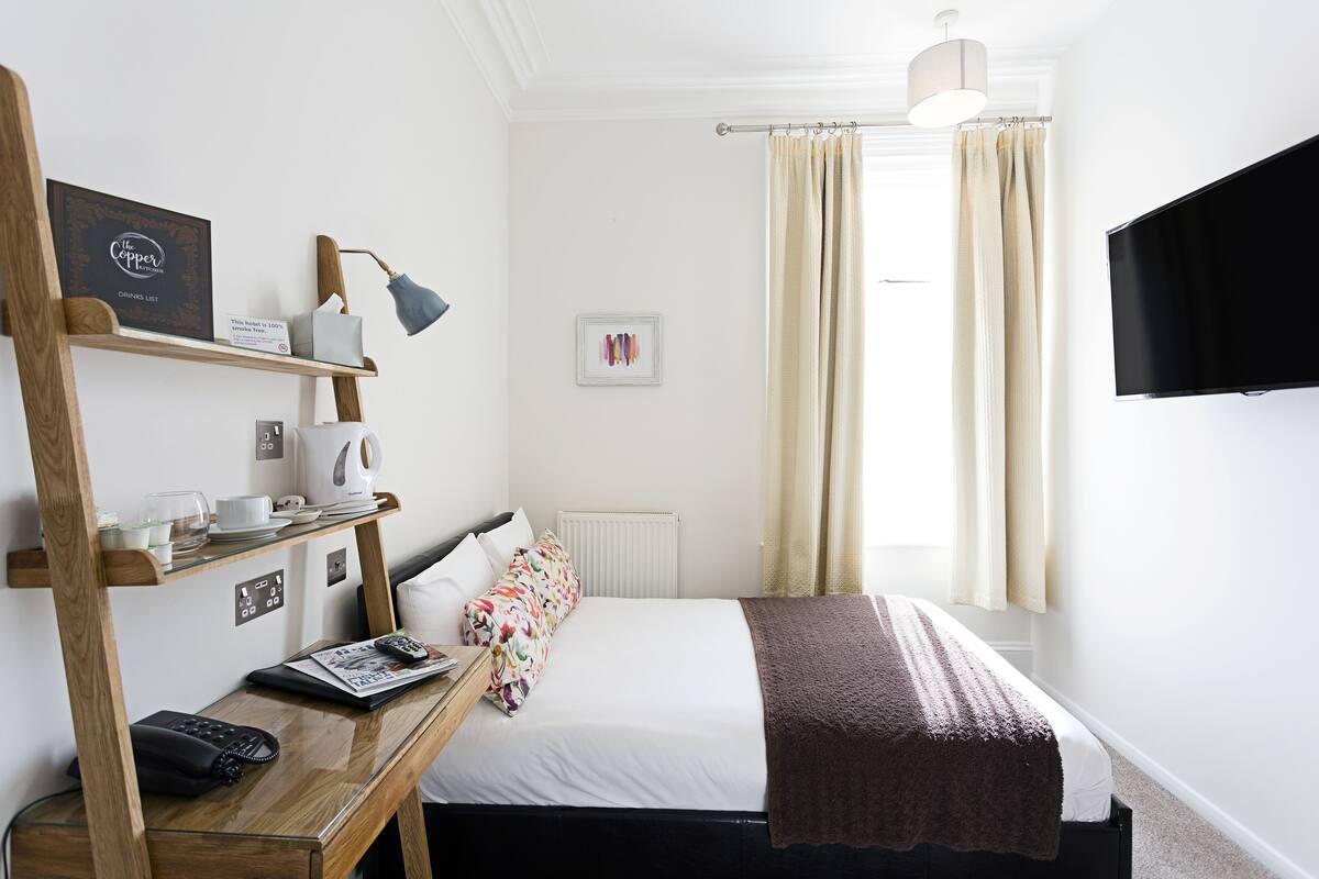 Een hotelkamer met een bed, nachtkastje met lamp, televisie en raam met gordijnen.