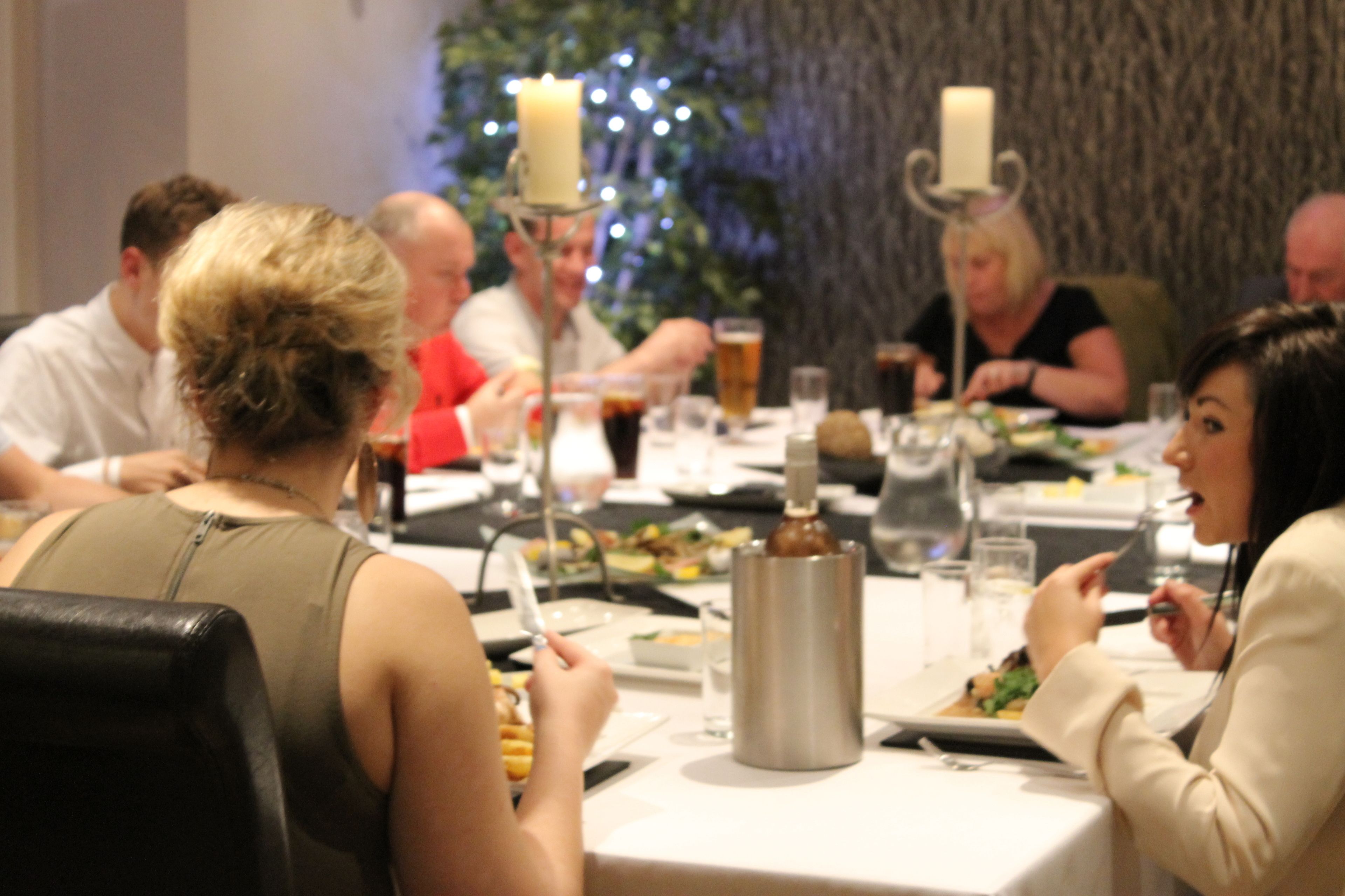 Um grupo de pessoas jantando à mesa, com um candelabro, um jarro de metal e um copo de cerveja.