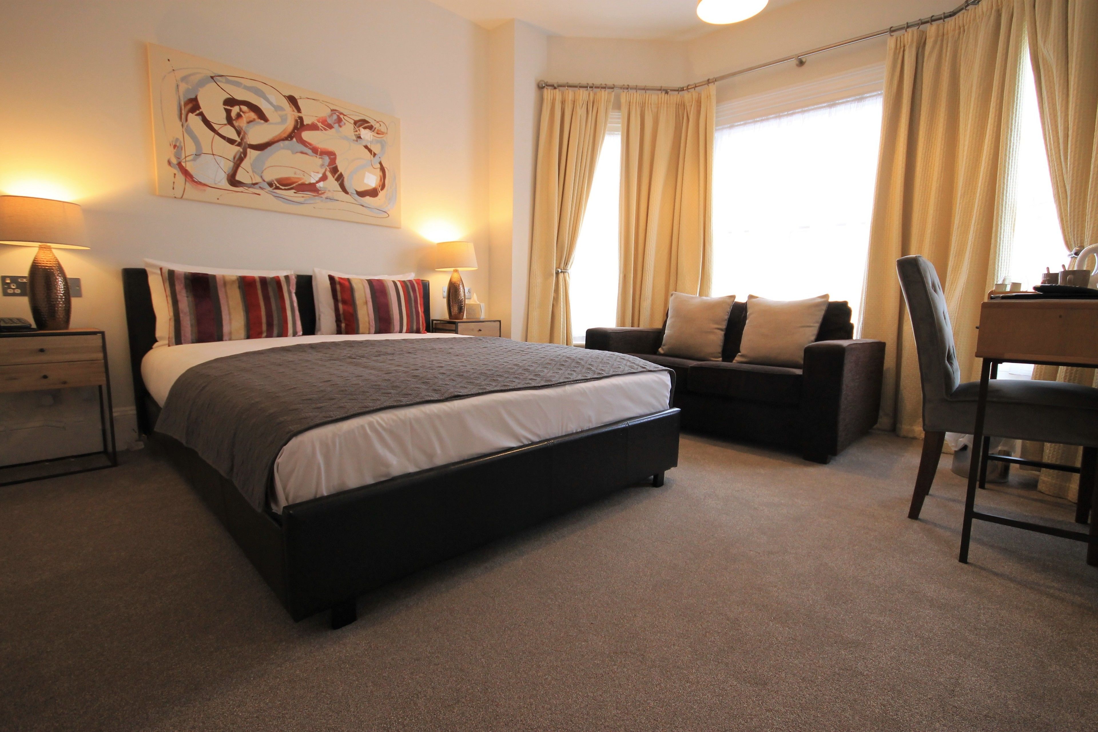A bedroom with a large bed, a desk, and a chair. There is a window with curtains, a lamp, and a painting on the wall.