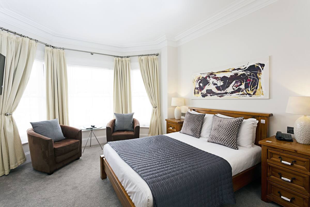 Een hotelkamer met een groot bed, twee fauteuils, een nachtkastje met een lamp en een kunstwerk aan de muur.