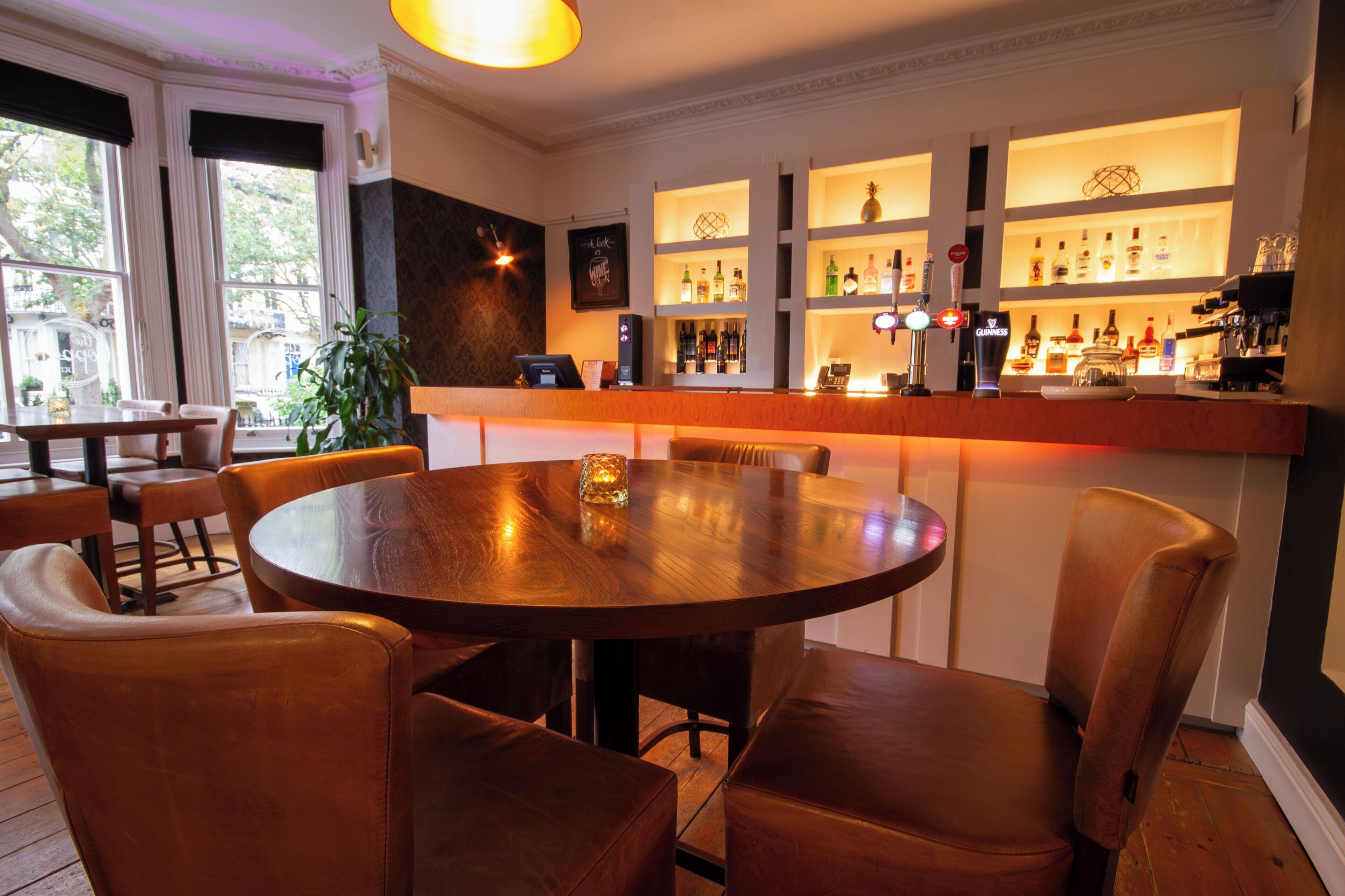 Uma área de bar com mesa de madeira redonda e cadeiras de couro, prateleiras com garrafas e uma vista através de grandes janelas.