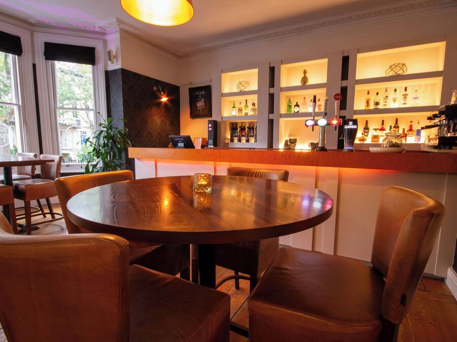 Uma área de bar com mesa de madeira redonda e cadeiras de couro, prateleiras com garrafas e uma vista através de grandes janelas.
