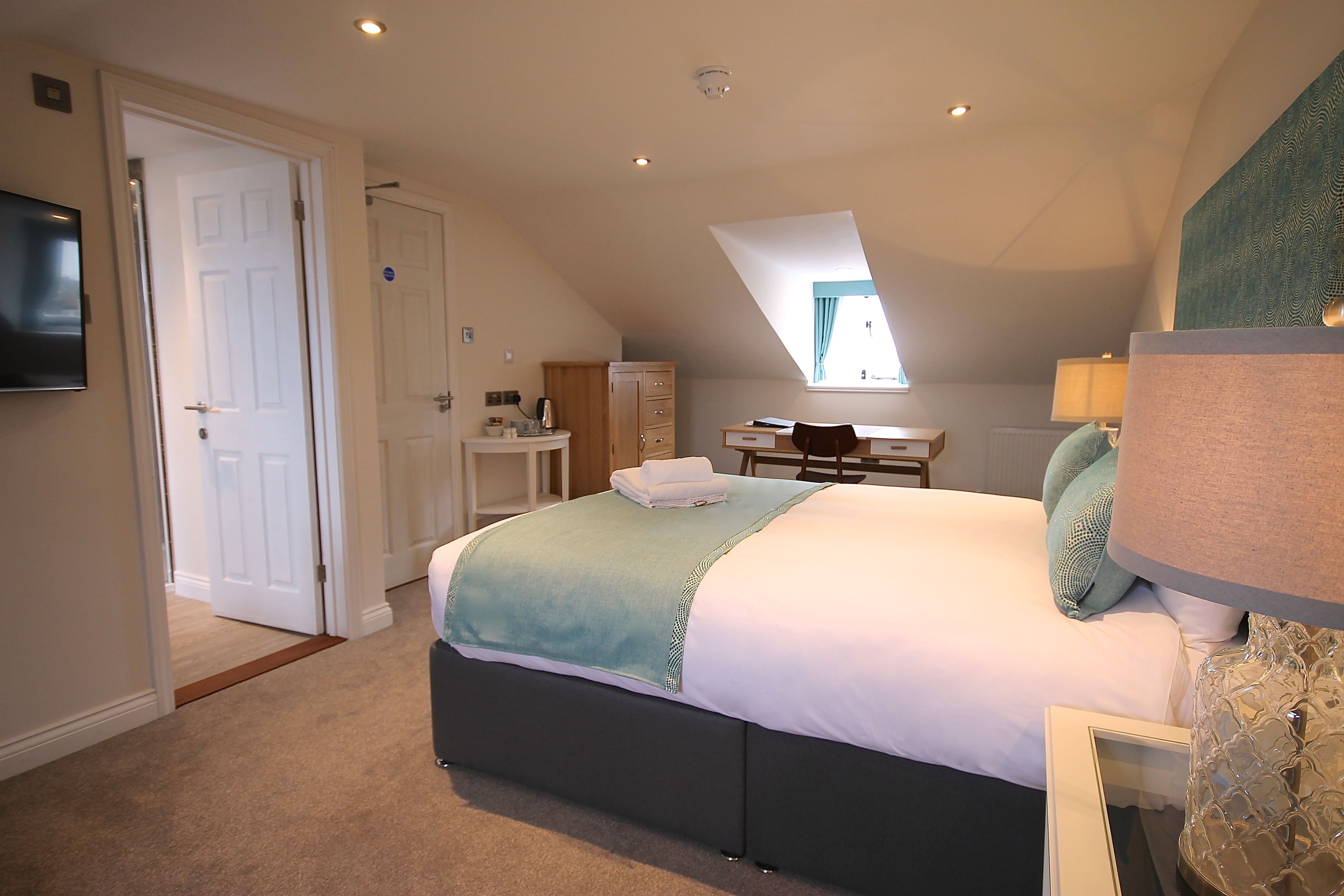 A bedroom with a bed, a desk, and a chair. There is a door leading to another room and a window providing natural light.