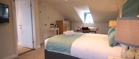 A bedroom with a bed, a desk, and a chair. There is a door leading to another room and a window providing natural light.