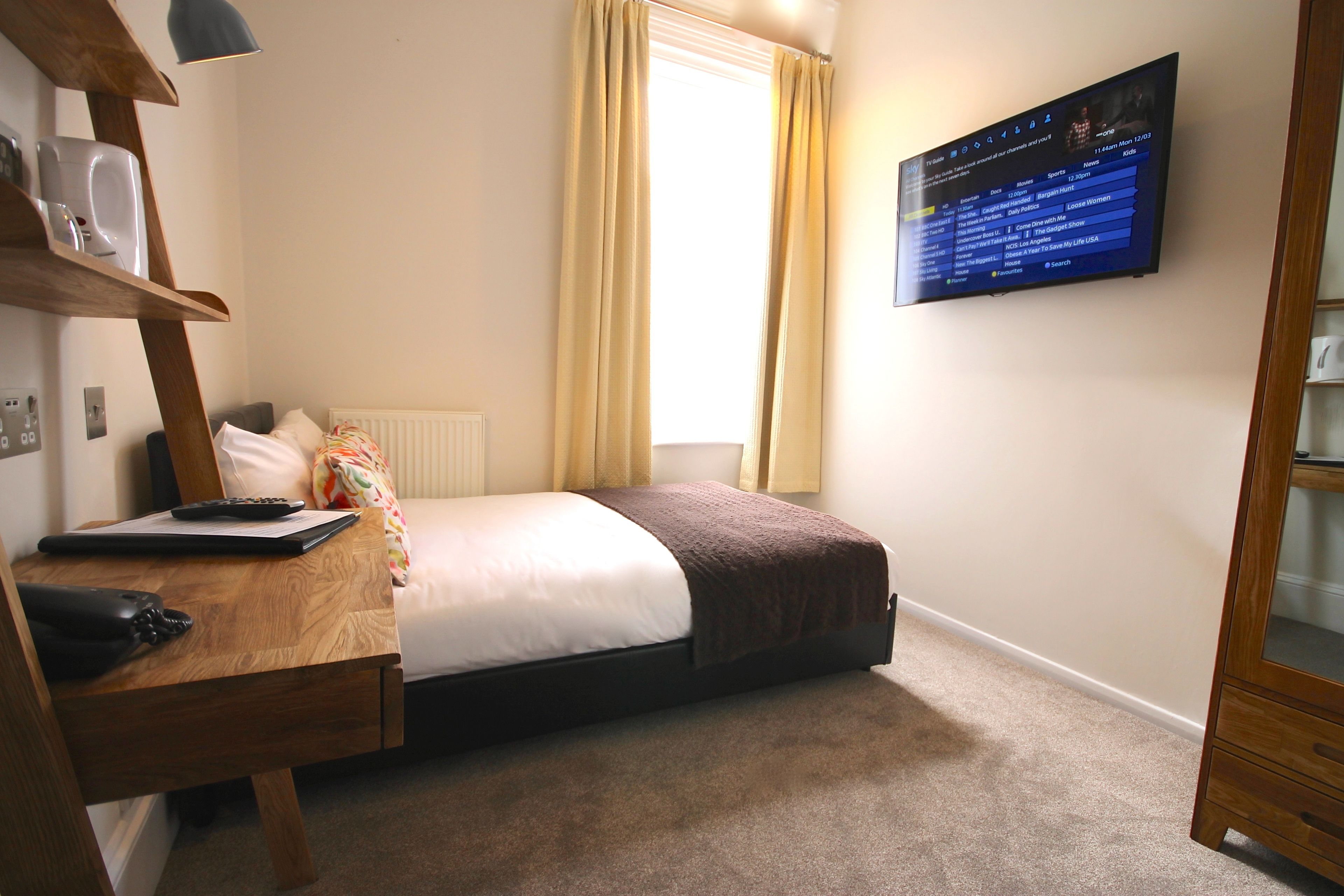 A bedroom with a bed, a desk with a computer, a telephone, and a TV mounted on the wall.