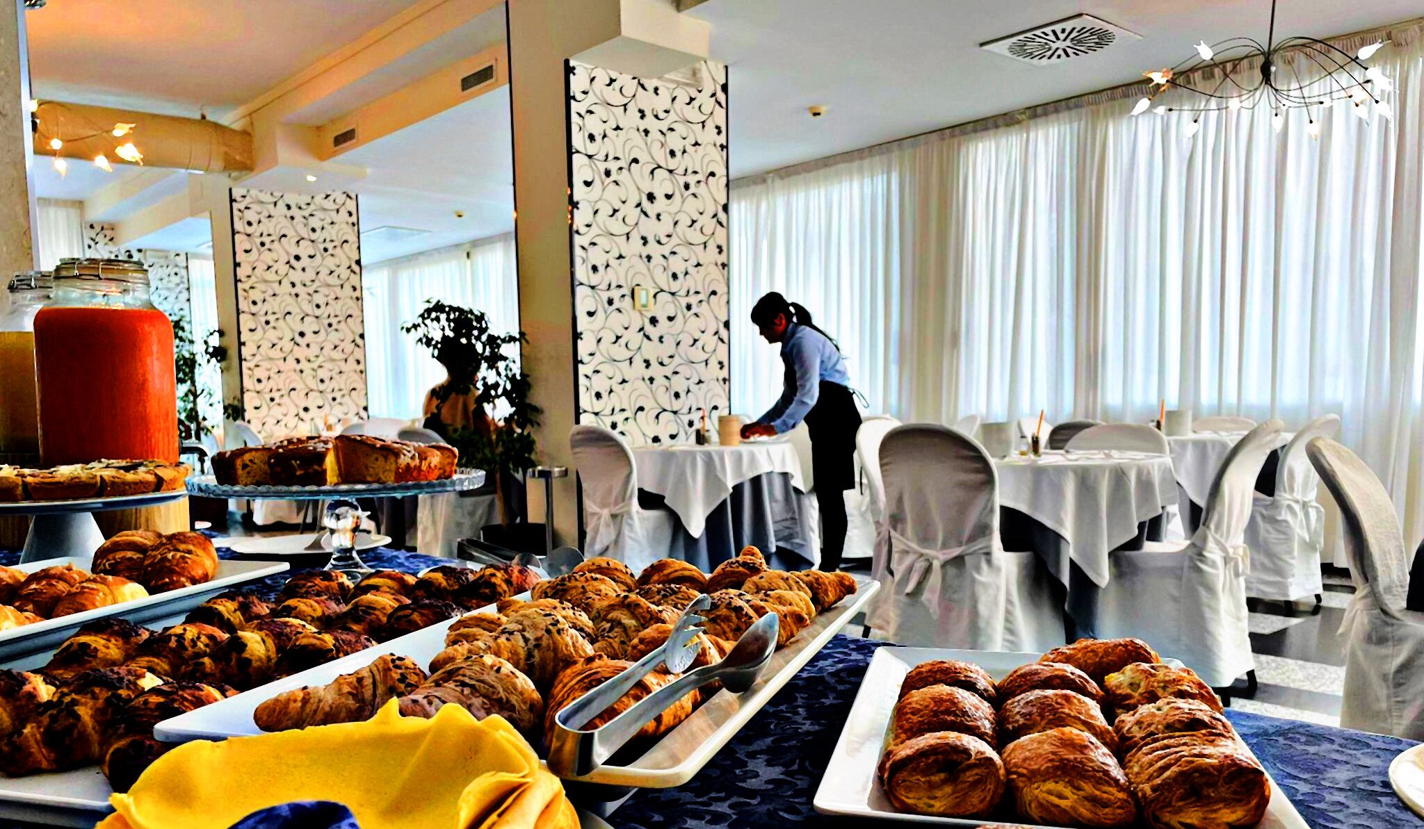 Montaje de bufé con diversos pasteles y un pastel, una persona sirviendo comida, y una sala con candelabro y ventanas amplias.