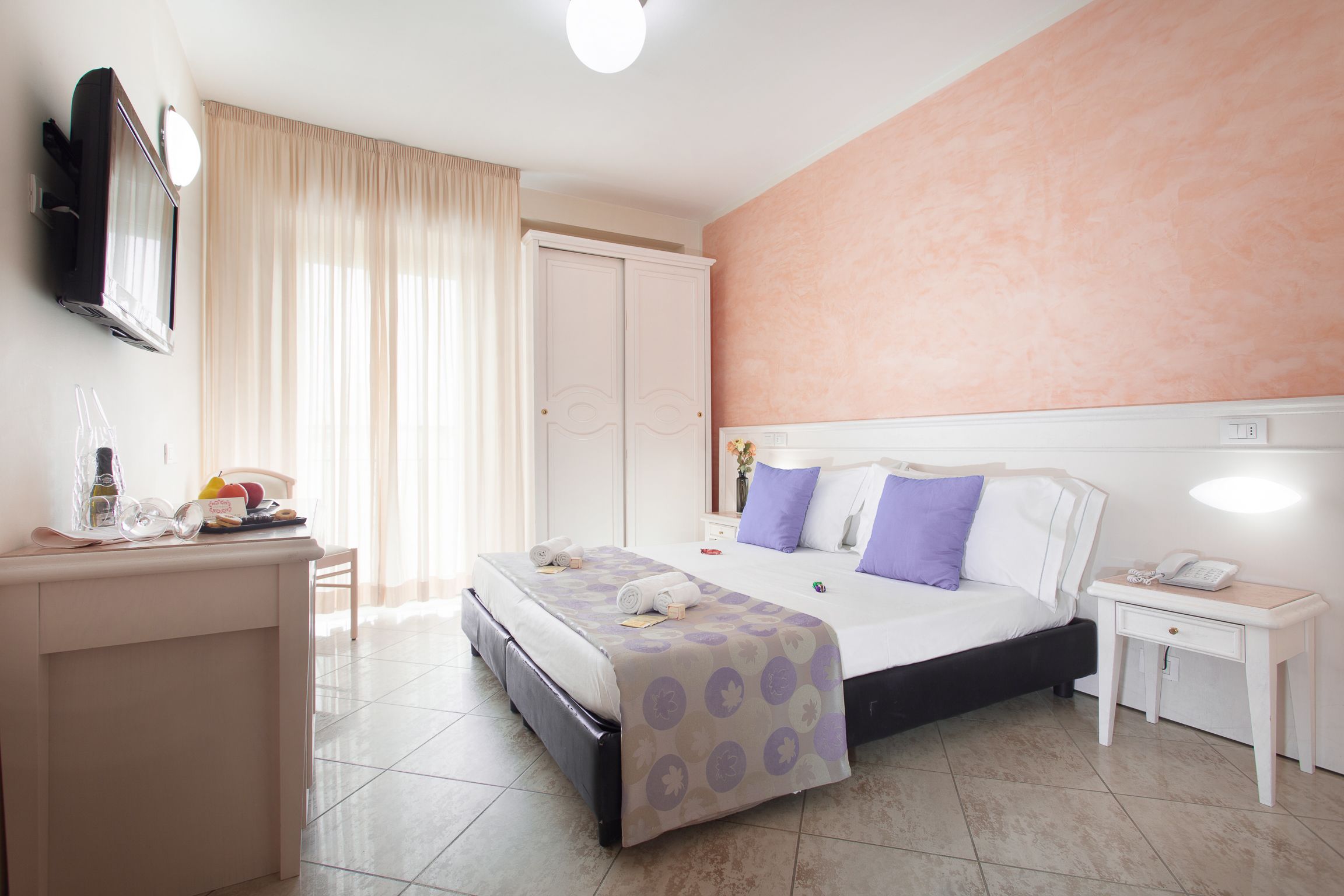 A hotel room with a bed, bedside table, and a desk with a mirror and toiletries.