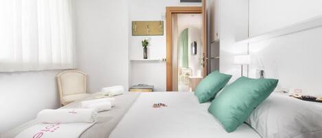 A hotel room with a neatly made bed, a chair, a bedside table with a lamp, and a view of the hallway.