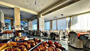A hotel dining area with tables set for breakfast, a buffet with pastries, and a large window with curtains. - Hotel Ambra (Cervia)