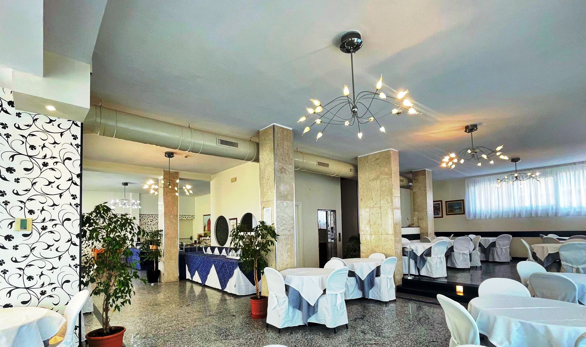 A spacious indoor venue with multiple tables and chairs, decorative lighting fixtures, and potted plants.
