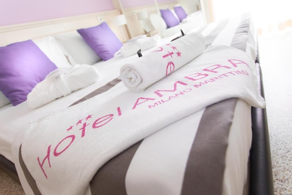 Una cama de hotel con almohada a rayas blancas y grises y una toalla blanca con el bordado 'Yotei Ambra Milano Marittima'.