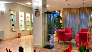 A spacious lobby with a glass enclosure, red armchairs, and a potted plant. - Hotel Ambra (Cervia)