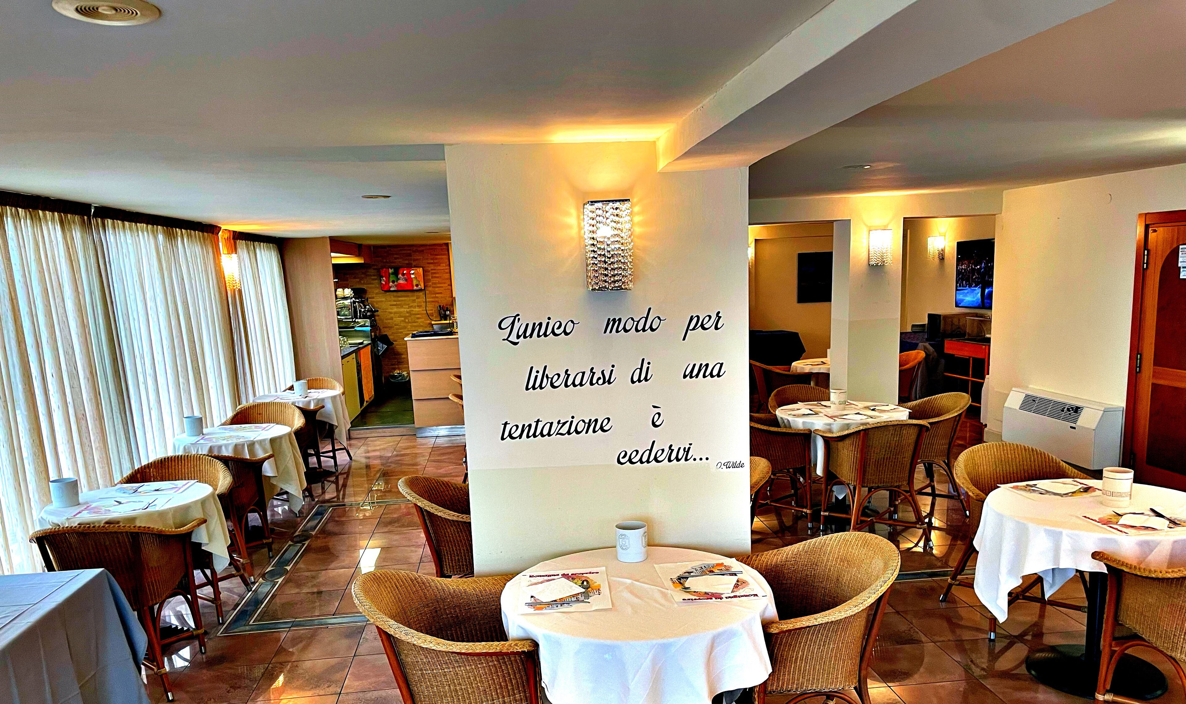 Ein Restaurant mit Korbstühlen und Tischen, im Hintergrund eine Küche und ein Zitat an der Wand.