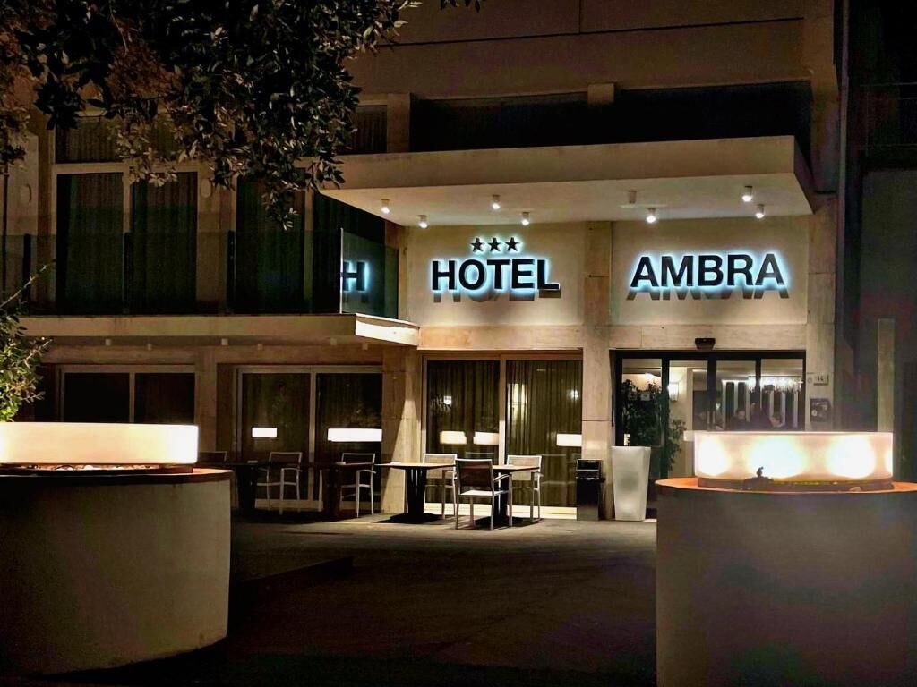 Entrada de un hotel con un letrero que dice "HOTEL AMBRA" y un área exterior bien iluminada con mesas y sillas.