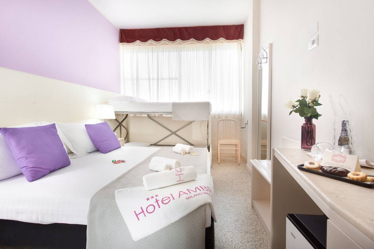 A hotel room with a bed, a desk, and a chair. There are towels and a tray of donuts on the desk.