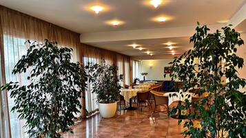 Uma sala de jantar de hotel com várias mesas e cadeiras, grandes vasos de plantas e piso de azulejos.